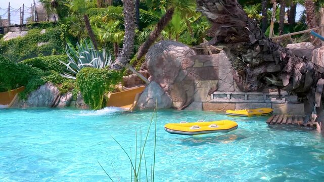 Inflatable yellow rafts gently bob in the crystal-clear blue water at the water park, surrounded by palm trees. The exotic scenery creates the relaxing atmosphere of a summer resort.