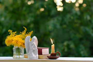 Obraz premium Beautiful composition with angel figurine, candle in candlestick, bible books, bouquet of yellow flowers in glass vase on table outdoor, nature background. symbol of faith, pray, believe, christianity