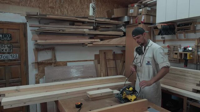 Authentic local cabinetmaker setting up handheld circular saw and unwinding power cord on timber stack at workbench in bright garage studio preparation b-roll