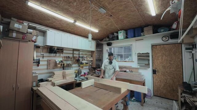 Authentic male carpenter lifting a heavy long pine board onto a table saw sled inside a cluttered domestic garage workshop wide angle establishing shot preparing for manual woodworking process