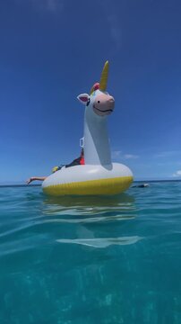 Child perched on inflatable unicorn, small silhouette against vast blue sky, toes dangling, lifevest visible, playful solo moment and confident balance on calm sea, vertical video