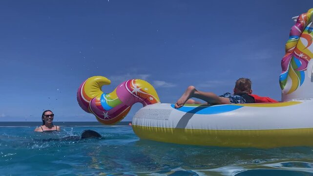 Two caucasian boys on inflatable unicorn pool float, mother guiding and laughing, bright blue water under clear sky, rainbow mane and colorful tail, playful splashes, sunlit family