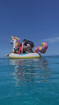 Mother and kids balancing on unicorn float in ocean, playful tumble and laughter, colorful tail and splash, swimsuit closeup, bonding moment under warm sunshine, vertical video