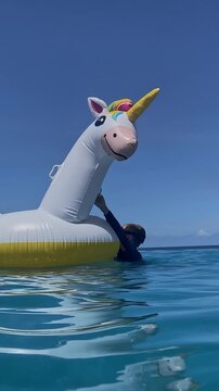 Unicorn float bobbing on turquoise sea, two boys balancing and laughing while gripping rim, snorkeler mask visible, clear water shimmer and gentle drift, wide perspective capturing