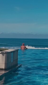 Man In Infinity Pool Bali Sea Horizon, Traveler Relaxing On Pool Edge With Back To Camera, Lifebuoy Mounted On Palm Column, Clear Blue Water Blending With Ocean, Bright Sunlight, Calm Waves, Resort