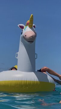 Mother and kids balancing on unicorn float in ocean, playful tumble and laughter, colorful tail and splash, swimsuit closeup, bonding moment under warm sunshine, vertical video