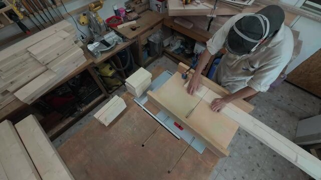 Authentic unrecognizable woodworker cutting pine timber into small blanks on a table saw inside a cluttered garage workshop overhead top down handheld shot of repetitive batch production process