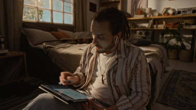 Young Caucasian man sitting on floor, using stylus to draw on tablet screen while wearing wireless earbuds and enjoying inspiring music. Showcasing remote work, digital art, or online learning