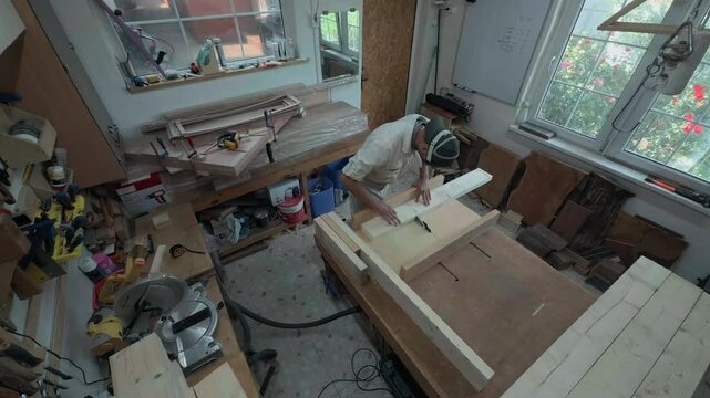 Professional woodworker wearing respirator cutting timber boards on a table saw machine inside domestic garage workshop high angle top down wide shot of authentic local manufacturing process
