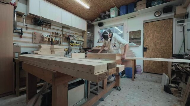 Self-employed woodworker in protective wear sorting rough timber planks on large workbench inside domestic carpentry shop handheld waist level shot of authentic small business owner working