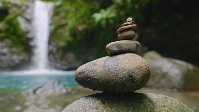 Balanced zen stone cairn stacked on mossy river boulder with turquoise waterfall pool and lush green tropical jungle foliage in soft focus background. Peaceful meditation scene.