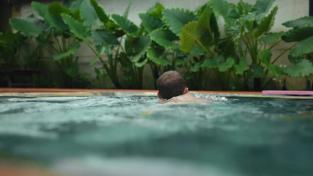 A man swims in a turquoise pool surrounded by lush tropical foliage. Water ripples and splashes as he moves through the calm pool with large green elephant ear plants behind.