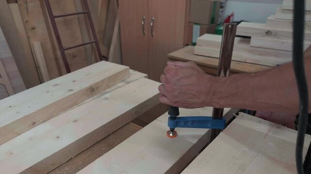 Unrecognizable male carpenter fixing wooden pine boards with a blue metal clamp on a workbench in an authentic carpentry workshop close up shot of manual labor hand holding tool securing the wood