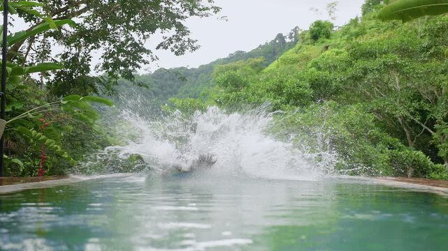 Slow motion footage of father and son jumping together into an infinity pool surrounded by lush tropical rainforest and misty green mountains creating a massive water splash.