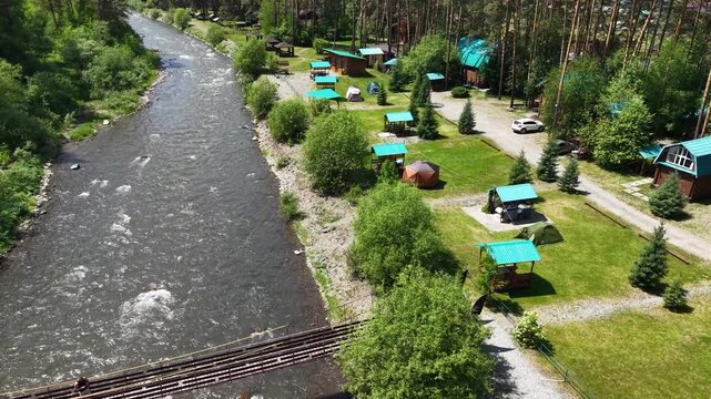 A cinematic high-altitude drone aerial shot flying forward over a beautiful riverside camping resort with cozy barbecue gazebos and green lawns near Kamlak in Altai.