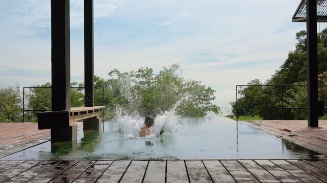 Silhouetted child runs across wet wooden deck and leaps into a stunning infinity pool at a luxury tropical resort, creating a dramatic splash against lush green trees and overcast sky backdrop.