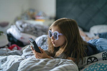Child with sunglasses playing video game on hand-held device in bed © Miljan Živković