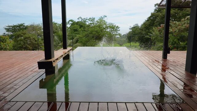 Slow motion footage of a young boy jumping into a modern infinity pool surrounded by lush tropical jungle. Wooden deck and contemporary architecture frame scenic rainforest vista under cloudy skies.