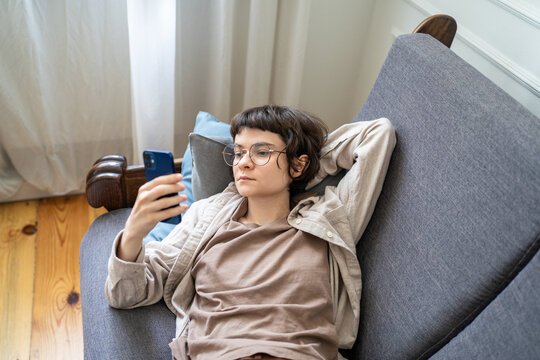 Lazy woman browsing social media on smartphone while lounging on sofa. Zoomer girl wasted time. Device dependency, endless scrolling habit, modern leisure, lonely isolation. Phone addiction concept