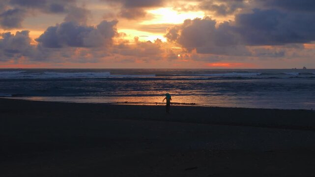Lone child stands silhouetted against a vibrant ocean sunset on a sandy beach. Dramatic clouds glow with orange and golden light as waves roll onto shore. Peaceful coastal evening scene.
