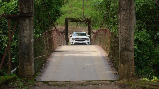 White pickup truck crosses a rustic suspension bridge with weathered concrete pillars and red mesh railings, surrounded by dense tropical jungle foliage under overcast skies in a remote area.