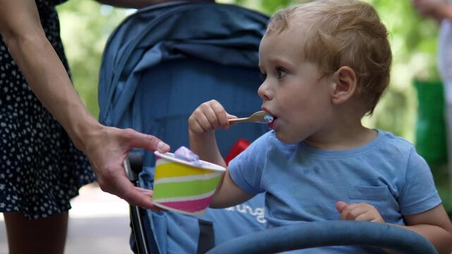 Chronological family scene: toddler seated right foreground in blue stroller is fed frozen dessert by adult whose hands enter from left foreground holding