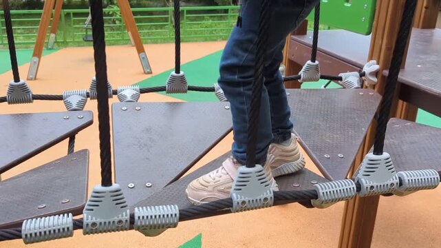 balancing child step across textured, triangular platform steps on a modern playground bridge structure supported by thick black vertical ropes.