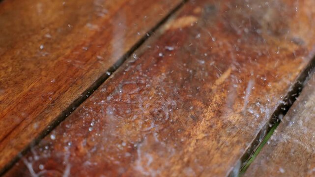 Slow motion close up of raindrops hitting wet wooden deck planks creating splashes and ripples. Water droplets bounce off the rich brown stained wood surface during rainfall.