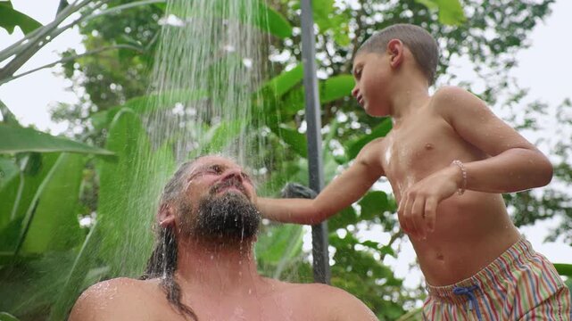 Father and son share a playful moment under an outdoor tropical shower as water streams down. Young boy touches dads wet hair amid vibrant green banana leaves and lush garden foliage on sunny day.