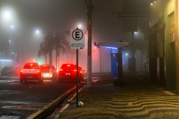 Naklejka premium A car drives through thick fog on an urban street under traffic lights at night in downtown Marilia, Sao Paulo state, 