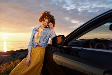 Fototapeta premium A woman with curly hair rests against a sleek car at sunset, wearing a striped tee and denim jacket, showcasing a casual, carefree lifestyle and travel vibe.