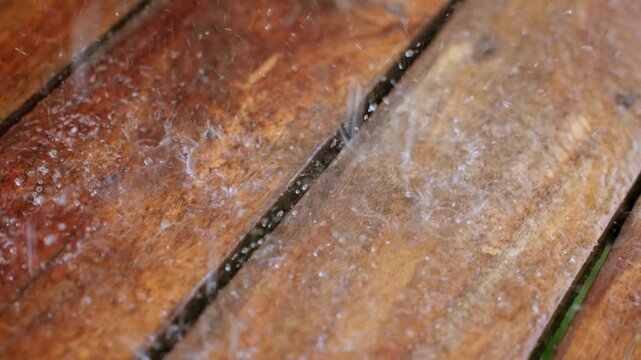 Slow motion close up of raindrops falling and splashing on weathered wooden deck planks. Water droplets create dynamic splash patterns on the warm brown timber surface with visible wood grain texture.