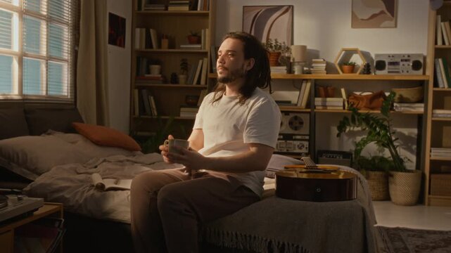 Caucasian young adult man with dreadlocks sitting on bed in home studio, drinking coffee and looking thoughtful. Video ideal for mental health awareness or remote work balance campaigns