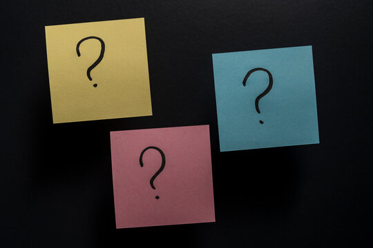 Yellow pink and blue post-it papers with handwritten question marks on dark surface. Minimalist concept for customer support FAQ pages choices brainstorm and decision making