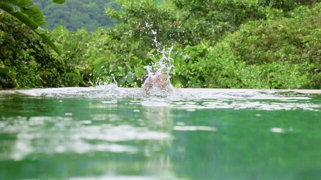 Young boy swimming through calm turquoise waters of a luxurious infinity pool with water splashing around him, surrounded by dense tropical rainforest vegetation and misty mountain backdrop.