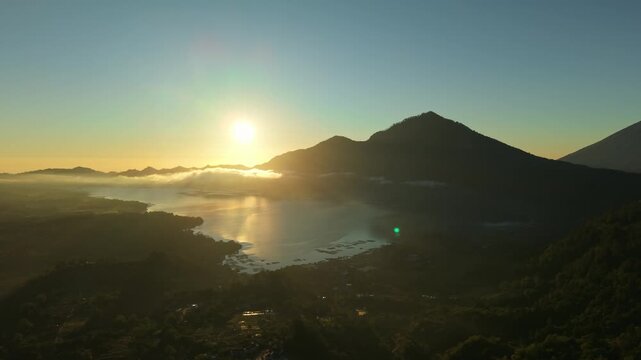 Aerial view of Lake Batur and Mount Abang at sunrise with golden light reflecting on the water surface and mist over the volcanic landscape in Kintamani, Bali, Indonesia.
