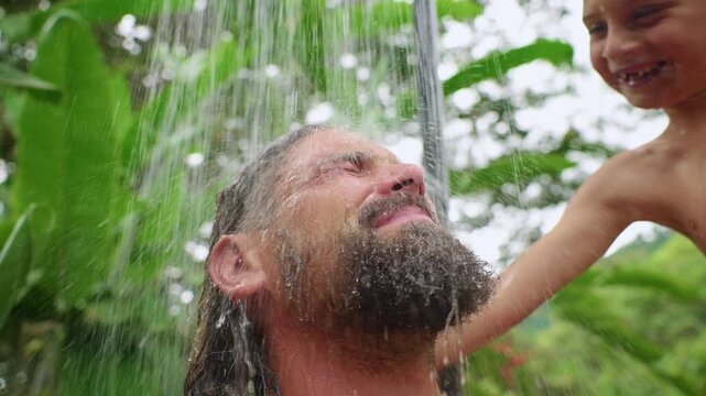 Son helps wash his father head under refreshing outdoor shower surrounded by lush tropical banana leaves. Water droplets cascade in slow motion as bearded man enjoys bonding moment with child.