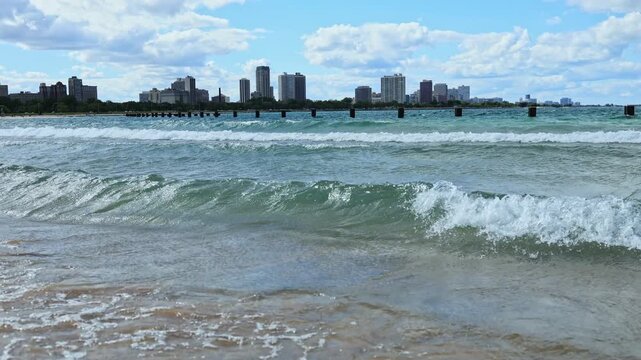 People walk along the shore while the water laps at the sand. The city skyline stands in the background under a blue sky with clouds.
