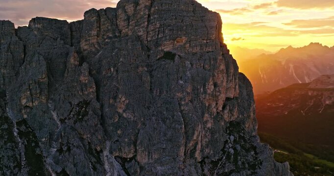 Smooth aerial flight along towering Dolomite cliffs during sunset. Golden sunlight creates a dramatic sunstar effect, illuminating rugged rock formations and alpine landscapes in Italy.