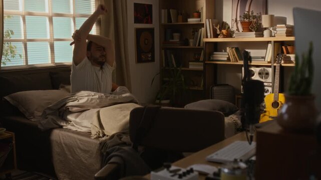 Young Caucasian man waking up and stretching in bed inside home studio apartment. Clip is ideal for commercials promoting remote work lifestyle, freelance routines, and morning habits