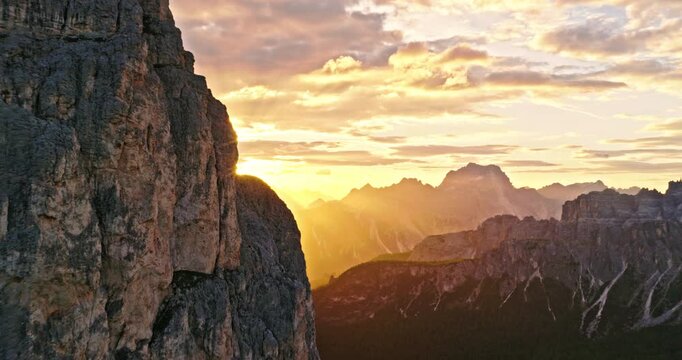 Smooth aerial flight along towering Dolomite cliffs during sunset. Golden sunlight creates a dramatic sunstar effect, illuminating rugged rock formations and alpine landscapes in Italy.