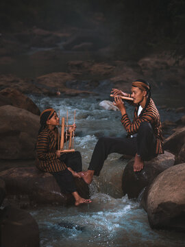 An Indonesian father and son in traditional Javanese surjan outfits play angklung and suling flute together by a rocky river.