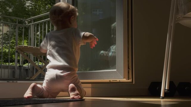 Curious baby exploring self-recognition while watching her reflection in a bright patio door on a sunny day.