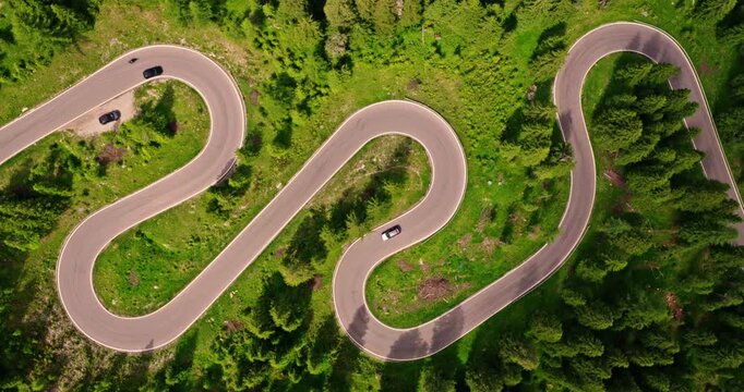 Aerial top down view of a winding mountain road through green forest landscape. Cars and motorcycles travel along dramatic hairpin turns, creating a scenic travel and transportation scene.
