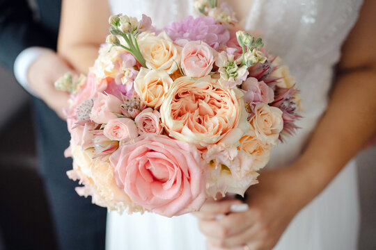 Young bride wearing a beautiful tulle white wedding gown while holding a lush bouquet of pink and cream roses, peonies and delicate seasonal flowers in a romantic pose holding arms with her groom