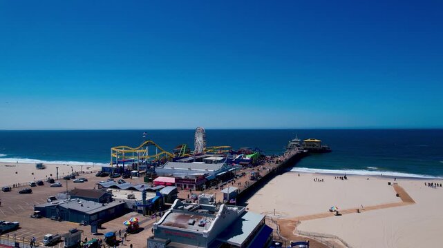 Aerial panoramic view of the iconic Santa Monica Pier in California featuring the Pacific Park amusement park, Ferris wheel, and roller coaster against a clear blue sky and ocean