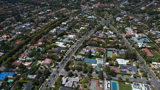 High-angle aerial view of a luxurious suburban neighborhood with sprawling mansions, private swimming pools, and tennis courts surrounded by lush green trees and palm-lined streets in California