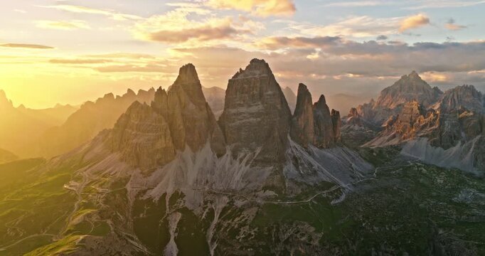 Cinematic drone view of Tre Cime di Lavaredo during golden hour. Warm sunlight bathes the iconic Dolomite peaks, creating a spectacular alpine landscape in northern Italy.
