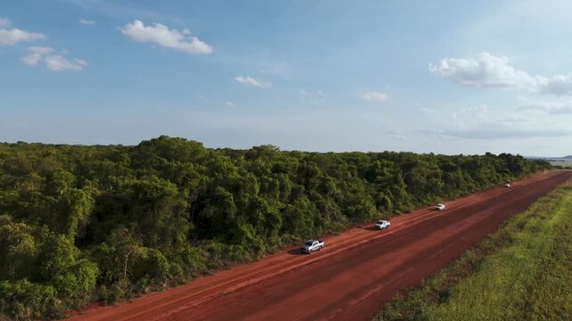 Aerial view of white vehicles driving on a long red dirt road through a lush green tropical forest under a clear blue sky with scattered clouds in a remote rural landscape