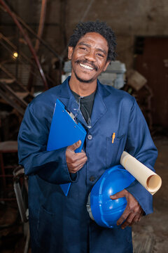 African worker in a factory holds a clipboard and a helmet while smiling at the camera